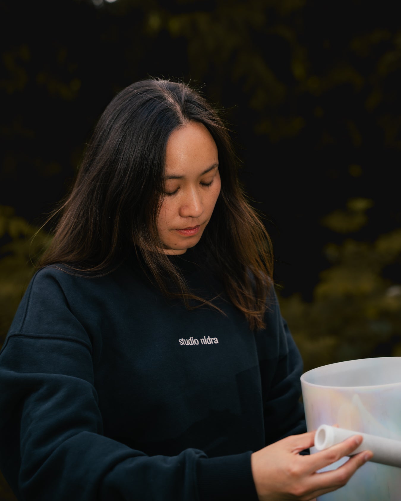Woman wearing The Crew black sweatshirt with Studio Nidra logo, holding crystal sound bowl.