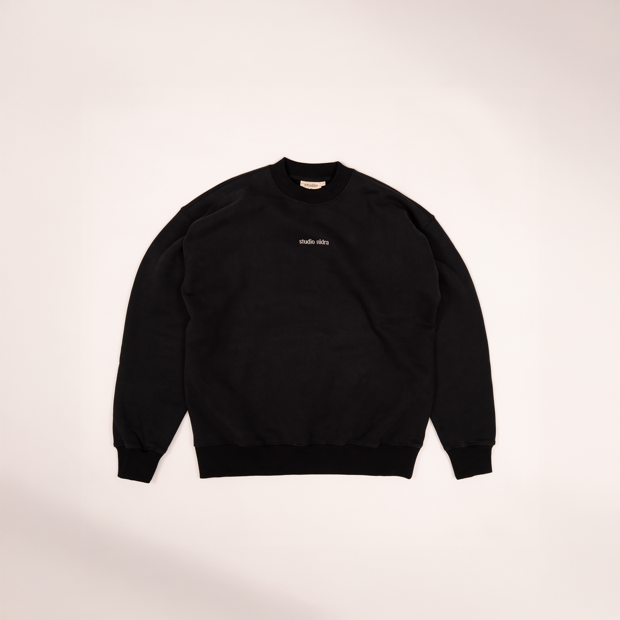 Flat lay image of the black crewneck sweatshirt with studio nidra logo embroidery on a light background