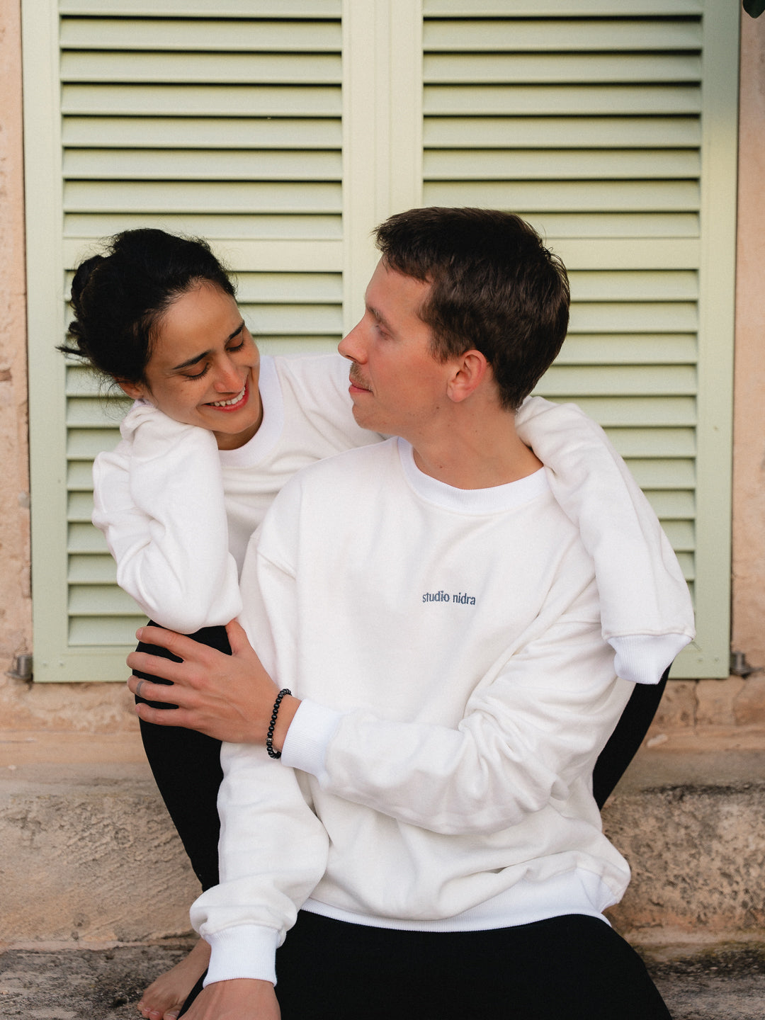 Two people in The Crew Sweatshirt limited edition in Marshmallow White sitting together in front of a building with green shutters.